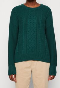 Pull en tricot bleu sarcelle avec un panneau central texturé, des poignets et un ourlet côtelés. Le tissu présente un mélange de motifs torsadés et de points.