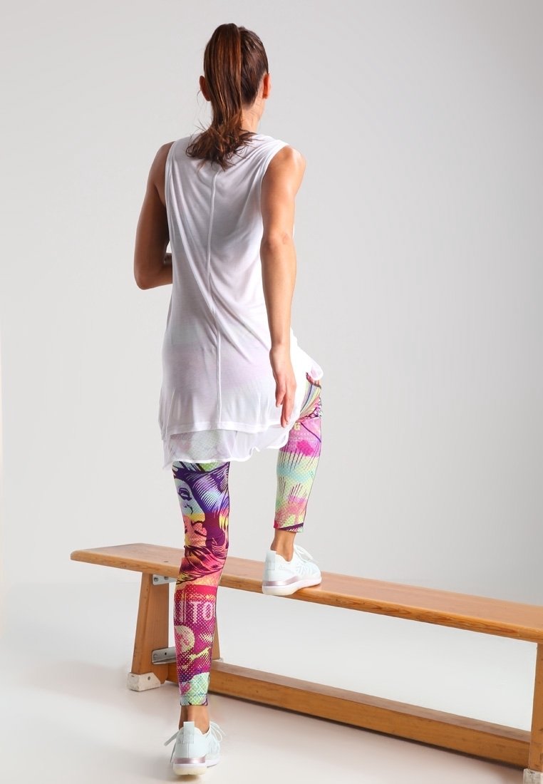Haut blanc sans manches, leggings colorés avec des motifs abstraits, et baskets légères. Personne debout sur un banc en bois.
