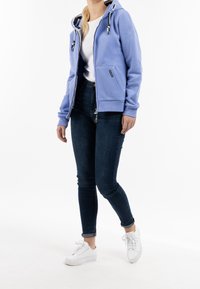 Felpa zip con cappuccio blu chiaro, dotata di cordino, tasca anteriore e dettaglio del logo, abbinata a jeans scuri e sneakers bianche.