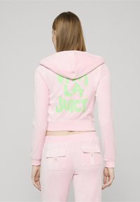 Kort rosa hoodie i mjukt material med en grön "VIVI LA JUICY"-grafik på ryggen. Har huva och ribbade muddar.