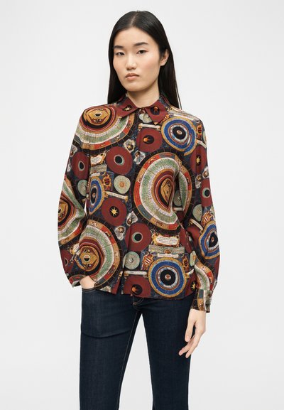 Γυναίκα που φορά μακρυμάνικο πουκάμισο με πολύχρωμα κυκλικά σχέδια και σκούρο τζιν, στέκεται μπροστά σε απλό φωτεινό φόντο.