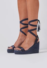Dunkelblaue Wedge-Sandalen mit Knöchelriemen. Glitzerndes Obermaterial und geflochtener Jutesohle. Offene Vorderseite mit glatter Textur.