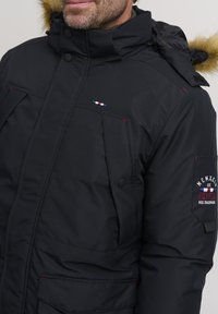 Black insulated parka with a fur-lined hood, featuring multiple pockets, red accents, and a logo patch on the sleeve. Water-resistant material.