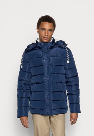 Navy blue puffer jacket with a high collar, snap buttons, and ribbed detailing. Features a detachable hood and contrasting white drawstrings.