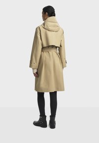 Kvinne iført en beige belted trenchcoat med hette, svarte leggings og svarte ankelstøvletter, står vendt bort fra kameraet mot en enkel bakgrunn.