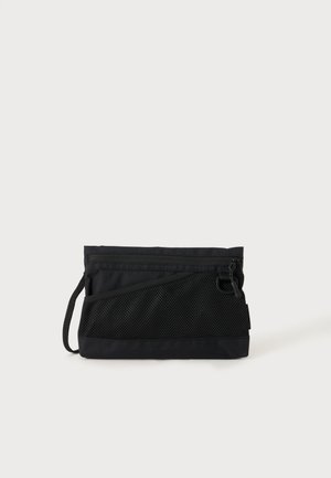 Pochette rectangulaire noire avec fermeture éclair en haut, poche avant en mesh et petite sangle sur fond blanc.