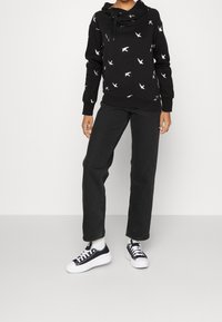 Sudadera negra con patrones de pájaros blancos, capucha con cordón, mangas largas y jeans rectos negros; combinada con zapatillas negras y blancas.