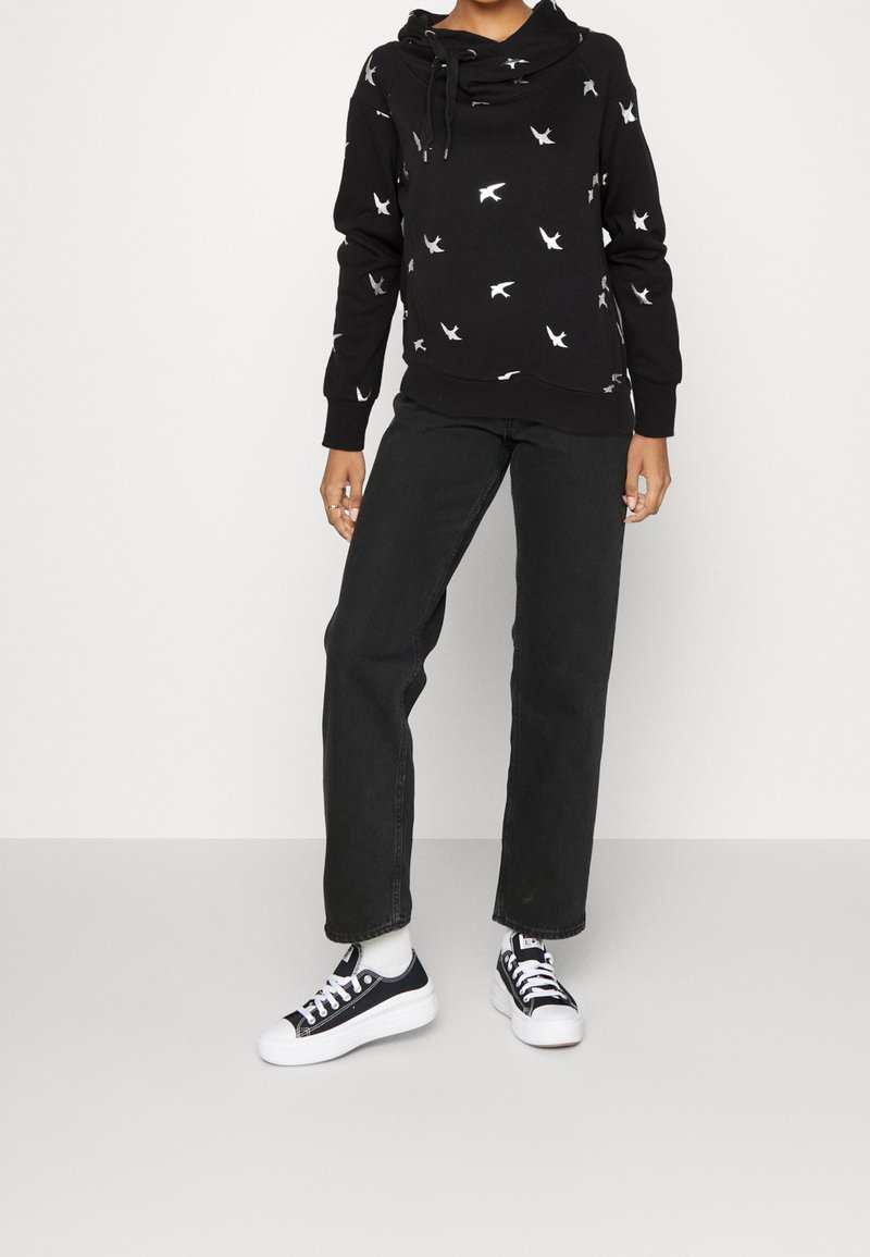 Sudadera negra con patrones de pájaros blancos, capucha con cordón, mangas largas y jeans rectos negros; combinada con zapatillas negras y blancas.