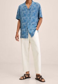 Kortebloedige, blauw gemusterde shirt met geometri5sche ontwerpen; gecombineerd met witte, relaxte broek en zwarte, gespen sandalen.