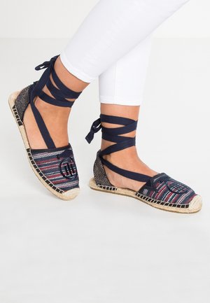 Pies con alpargatas azul marino de tela multicolor tejida y lazos para atar al tobillo, combinadas con pantalones blancos ajustados.