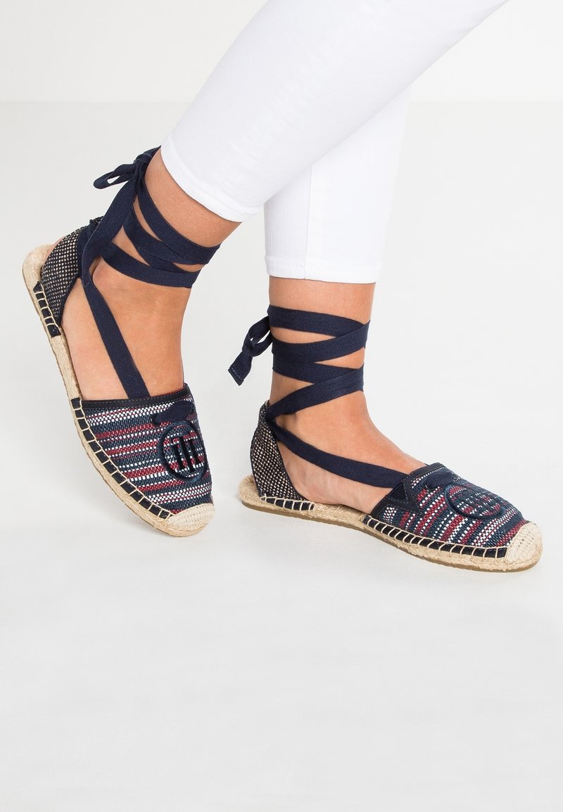 Pieds portant des espadrilles bleu marine avec un tissu multicolore tissé et des liens à nouer autour de la cheville, accompagnés d'un pantalon blanc ajusté.