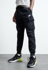 Sorte sweatpants med en afslappet pasform, der har sidelommer i cargo-stil og et hvidt Nike-logo. Kombineret med sorte og grønne træningssko.