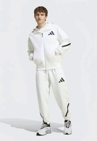 Balta sportinė treniruočių apranga su juodais akcentais; užtrauktuku užsegama gobtuvė su logotipu, prigludusios kelnės su juostelėmis ir sportiniai batai. Lygus tekstūra.
