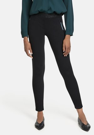 Personne portant un pantalon noir ajusté avec des poches latérales zippées, un chemisier vert foncé à manches longues, et des chaussures noires à bout pointu, debout.