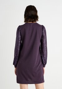 Lila klänning med kort ärm, med prydda puffärmar i paljetter. Tyget verkar mjukt med en slät yta, knälång design.