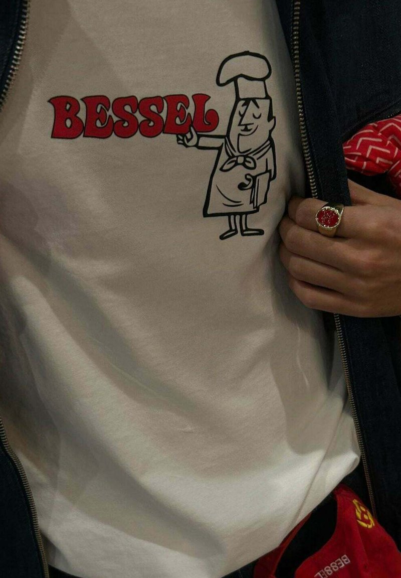 Camiseta de color crema con un gráfico de un chef de dibujos animados en contorno negro, con texto rojo que dice "BESSEL". Tela estampada visible en la cintura.