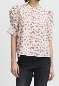 Květinová blůza s nafouknutými krátkými rukávy, světle krémovým pozadím a vzorem růžových květin. Obsahuje zapínání na knoflíky vpředu a uvolněný střih.