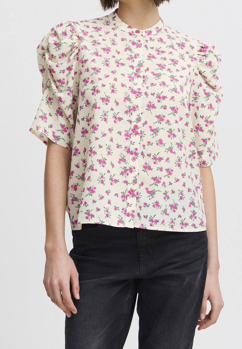 Květinová blůza s nafouknutými krátkými rukávy, světle krémovým pozadím a vzorem růžových květin. Obsahuje zapínání na knoflíky vpředu a uvolněný střih.