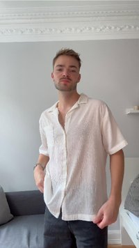 Camicia bianca a maniche corte con tessuto strutturato, taschino sul petto, e una vestibilità rilassata, abbinata a pantaloni scuri.