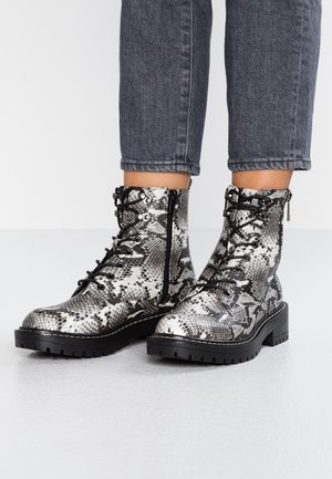 Bottes montantes à lacets avec un motif serpent noir et blanc, semelles épaisses noires, et fermetures éclair latérales, portées avec un jean court gris foncé.