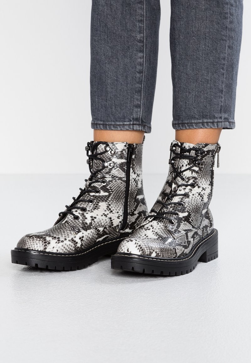 Bottes montantes à lacets avec un motif serpent noir et blanc, semelles épaisses noires, et fermetures éclair latérales, portées avec un jean court gris foncé.