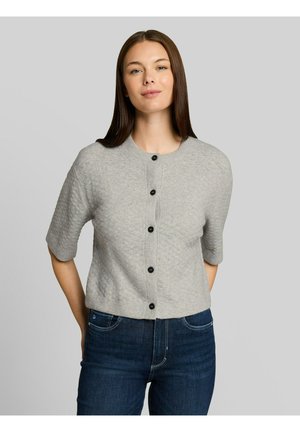 Femme aux cheveux bruns raides portant un cardigan gris à manches courtes boutonné et un jean bleu foncé, debout devant un fond neutre.