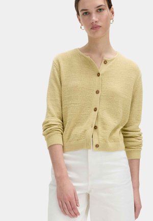 Vrouw draagt een lichtgele gebuttonde cardigan met lange mouwen en witte broek met hoge taille tegen een effen achtergrond.