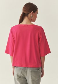 Haut rose à manches courtes avec une coupe décontractée et un décolleté carré, associé à un jean gris taille haute, montrant un design simple et épuré.