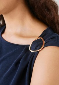 Navy blue top with soft fabric, featuring a gold oval accent on the shoulder and a draped design around the neckline.