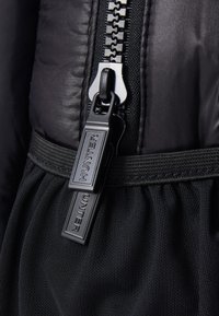 Detalle de una chaqueta de plumón negra que presenta una cremallera de alta resistencia, ribete elástico y un hardware prominente con la marca "HUNTER" en el tirador de la cremallera.