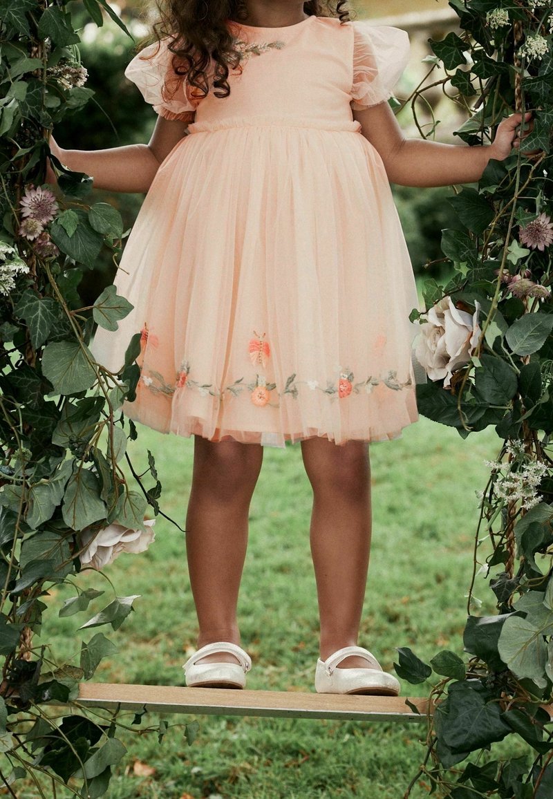 Enfant en robe pêche avec broderie florale se tient sur une balançoire en bois encadrée de feuilles vertes et de fleurs dans un jardin.