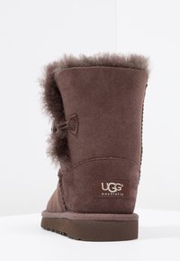 Brązowy zamszowy botek za kostkę z miękkim futerkiem, z akcentem w postaci guzika i teksturowaną podeszwą. Logo marki widoczne na tylnej części.