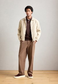 Homme debout les mains dans les poches, portant une veste beige, un polo marron, un pantalon marron ample et des baskets marron foncé avec des semelles blanches.
