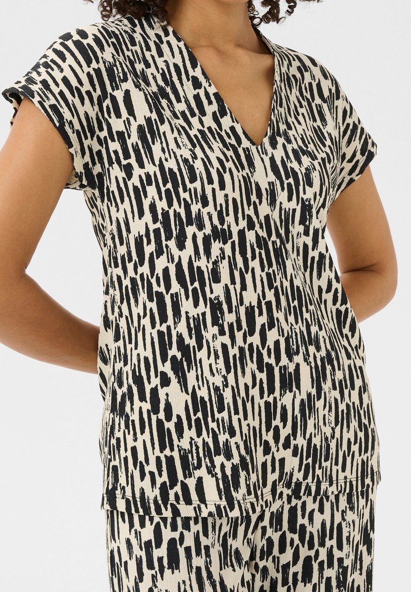 Femme portant un haut beige à manches courtes et un pantalon avec un motif irrégulier de coups de pinceau noirs verticaux sur un fond blanc uni.