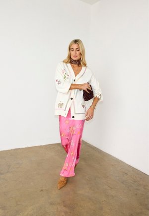 Femme debout portant un cardigan blanc brodé, une jupe longue à motifs roses, des chaussures marron pointues, tenant une pochette marron foncé contre un mur blanc uni.