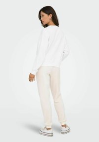 Felpa bianca in cotone con collo rotondo, abbinata a pantaloni beige chiaro. Il modello indossa sneakers bianche, mostrando un taglio rilassato e un design semplice.