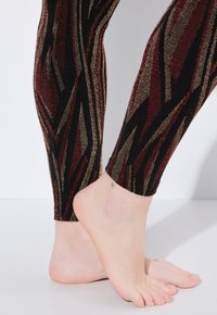 Leggings neri e oro con un motivo geometrico e dettagli in rosso e metallici. Inquadratura ravvicinata delle caviglie e dei piedi nudi. Texture liscia.