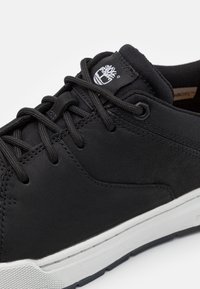 Gros plan d'une basket noire avec des lacets noirs, une languette en mesh, une semelle blanche et un logo arbre blanc sur la languette.