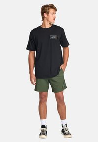 T-shirt nera con una grafica colorata di montagne sul petto, abbinata a pantaloni corti verdi con un motivo a logo. Sneakers nere completano l'outfit.