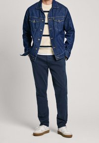 Jeansjacka med två bröstfickor, vit tröja med rund hals och mörka ränder, marinblå chinos och vita sneakers med bruna sulor och detaljer.