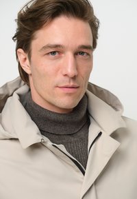 Beige vattentät jacka med huva, med en svart dragkedja, lager över en mörkbrun stickad polotröja, slät textur och skräddarsydd passform.