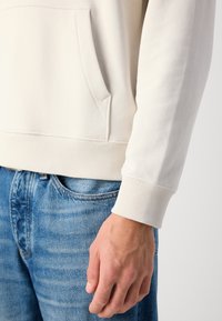 Hellbeige Kapuzenpullover mit Kängurutasche, geripptem Saum und Bündchen, getragen über blauen Jeans. Weiche Textur, lässiges Design.