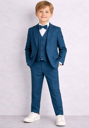 Jeune garçon debout, portant un costume bleu avec gilet, chemise blanche, nœud papillon bleu et baskets blanches, souriant les mains dans les poches.