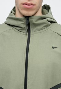 Jasnozielona kurtka Nike na zamek z kapturem, z czarnym zamkiem i logo, gładką teksturą oraz kontrastującymi czarnymi akcentami na dole.