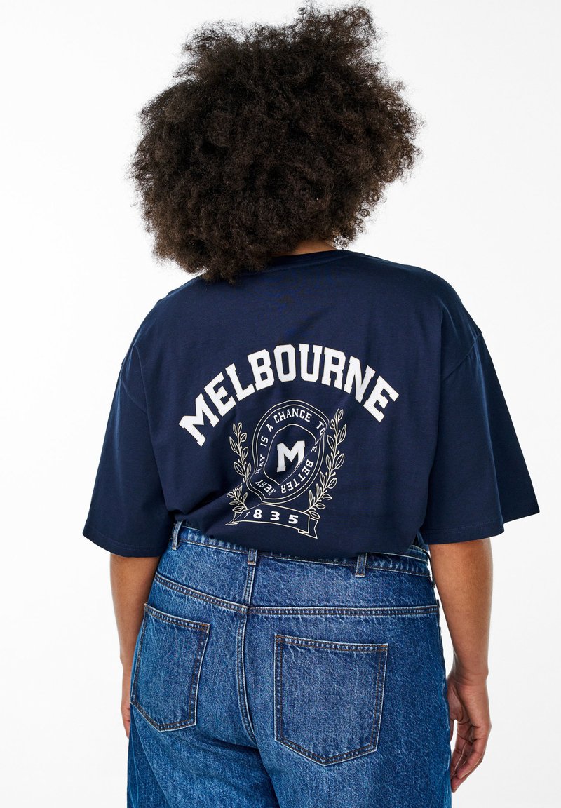 Námořnická modrá oversized tričko s nápisem "MELBOURNE" tištěným bílou barvou na zadní straně, s dekorativním emblémem a grafickým designem vavrínu.