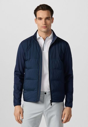 Jonge man die een marineblauwe doorgestikte jas draagt over een wit poloshirt en lichtgrijze broek, staand tegen een effen witte achtergrond.