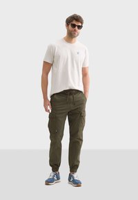 Witte T-shirt met blauw logo, olijfgroene cargobroek met elastische boorden en blauwe hardloopschoenen met witte veters en bruine zolen.