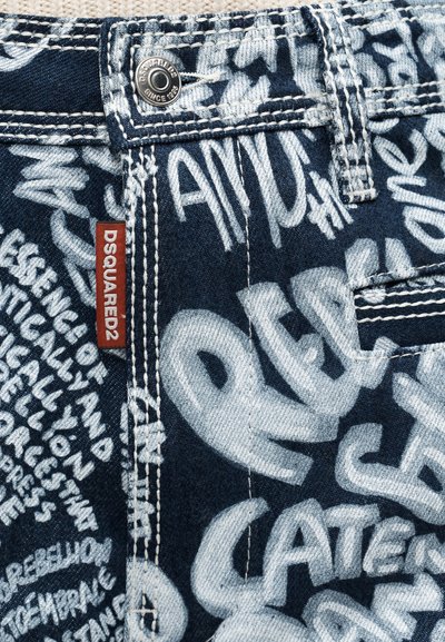 Ύφασμα από τζιν με σκούρο μπλε βάση που παρουσιάζει λευκό κείμενο σε στυλ γκράφιτι. Περιλαμβάνει κόκκινη υφασμένη ετικέτα και μεταλλικό κουμπί στη λεπτομέρεια.