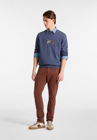 Granatowy sweter z haftem dachshunda, nałożony na jasnoniebieską koszulę z kołnierzykiem, w połączeniu z pomarańczowymi spodniami i beżowymi trampkami.