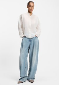 Weißes langärmeliges Hemd mit Knöpfen und Stehkragen; kombiniert mit weit geschnittenen, hellblauen Jeans und taupefarbenen spitzen Schuhen.